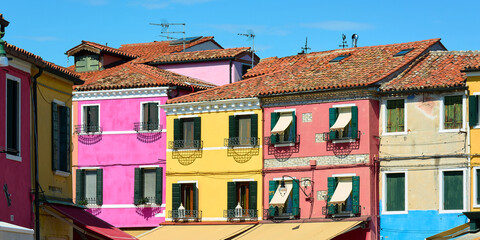 Horizontal panoramic view of facades of picturesque colorful houses against blue sky on sunny spring day in Burano, Italy.