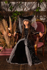 Art portrait of oriental mysterious girl Princess Queen dressed stylish costume, sitting on armchair in mystical dark room, looking at camera. Fabulous romantic image, fairy tale concept. Copy space