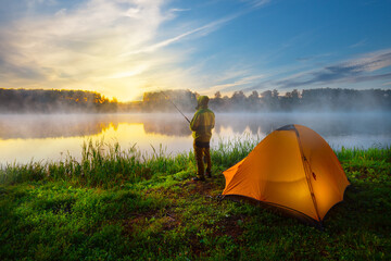 Fisherman on bank of foggy river near an orange tent in the early morning
