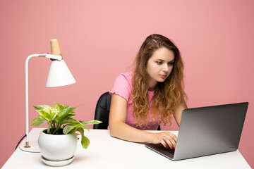 Cheerful young woman in a pink t-shirt using laptop computer while sitting at a desktop in a pink wall studio.