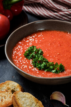 Gazpacho garnished with parsley in a plate with wavy edges. There is red pepper, tomato, garlic and  croutons on the table. Top view.