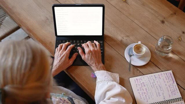 Point of view overhead closeup view of mature elderly woman hands typing on laptop tablet computer wearing ethnic clothes in a cafe coffee shop.