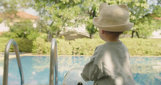 Happy little adorable Asian baby toddler girl in hat having good time in outdoor swimming pool sitting at poolside and looking at pure clear water in vacation holiday summertime slow motion