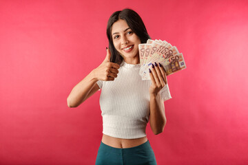 Happy brunette woman wearing casual top isolated over red background holding bunch of money and making okay gesture and looking to the camera with smiles. Win money prize lottery showing thumb-up.