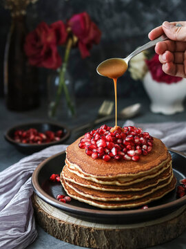 Stack of pancakes decorated with pomegranate, red flowers and pouring caramel on a black plate and a dark background