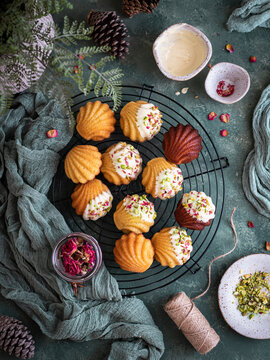 Festive madeleines on a cooling rack decorated with white chocolate, pistachios and red petals over a green background and cloth