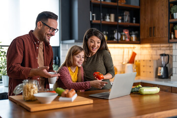 Family of tree is having fun while cooking together. They're all watching something funny on a laptop and laughing. Mom is holding fresh tomatoes, dad is holding a cup of coffee.