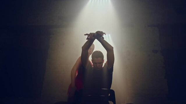 Front below view of man siting on chair in center,female dancer moves behind him in slow dance,they make hand movements in tango style,woman leaves frame,he rises to his full height,Loft atmosphere.