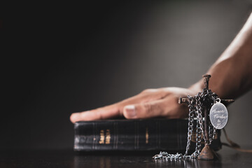 Mano en posición de orar o rezar y sosteniendo la santa biblia y cruza de clavos envuelto con un collar con placa que tiene grabada la frase “nunca pierdas la fe” en fondo negro y blanco y negro