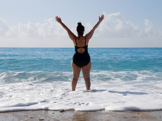 Fototapeta na wymiar a curvaceous woman in a black bathing suit stands in the sea, raising her hands
