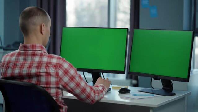 Young Man Working On Computer With Two Green Screen Mock Ups Sitting At Desk In Office