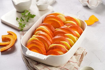Sliced and layered potato and pumpkin casserole in a white baking form on marble background, raw ingredients, oregano, whipped cream