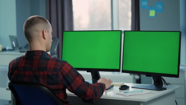 Young Man Working On Computer With Two Green Screen Mock Ups Sitting At Desk In Office. Office Worker Looks at Monitors And Writes Notes in Notebook