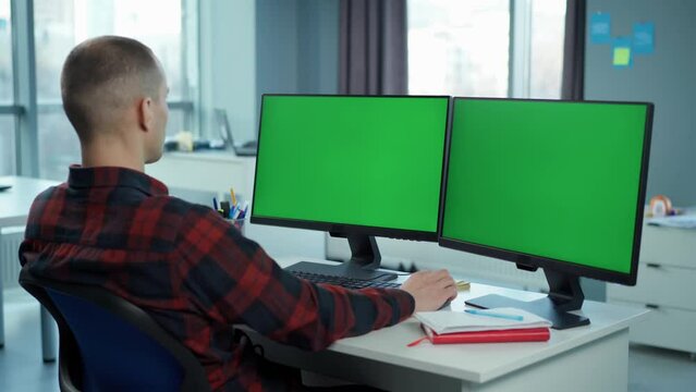 Young Man Working On Computer With Two Green Screen Mock Ups Sitting At Desk In Office