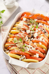 Sliced and layered potato and pumpkin casserole in a white baking form on marble background, raw ingredients, topped with roasted onions, feta cheese crumbs and whipped cream, ready to bake