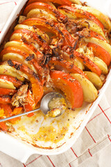 Sliced and layered potato and pumpkin casserole in a white baking form on marble background, topped with roasted onions, feta cheese crumbs and whipped cream