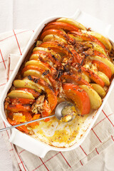 Sliced and layered potato and pumpkin casserole in a white baking form on marble background, topped with roasted onions, feta cheese crumbs and whipped cream
