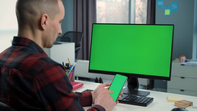 Man Uses Phone With Green Mock Up Screen on Background Green Screen Monitor in Office