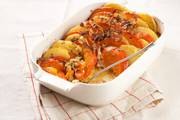 Sliced and layered potato and pumpkin casserole in a white baking form on marble background, topped with roasted onions, feta cheese crumbs and whipped cream