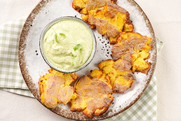 Trendy comfort snack crispy roasted crushed potatoes on a round plate with creamy avocado and garlic dip on a green checkered kitchen towel. top view