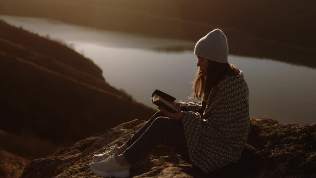 Young beautiful woman wrapped in a blanket reading the bible sitting on top of a rock with a beautiful view at sunset, admiring the view and nature, relaxing alone with thoughts