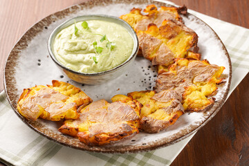 Trendy comfort snack crispy roasted crushed potatoes on a round plate with creamy avocado and garlic dip on a green checkered kitchen towel on wooden background