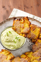Trendy comfort snack crispy roasted crushed potatoes on a round plate with creamy avocado and garlic dip on a green checkered kitchen towel on wooden background