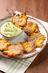 Trendy comfort snack crispy roasted crushed potatoes on a round plate with creamy avocado and garlic dip on a green checkered kitchen towel on wooden background
