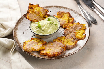 Trendy comfort snack crispy roasted crushed potatoes on a round plate with creamy avocado and garlic dip on a beige colored table cloth