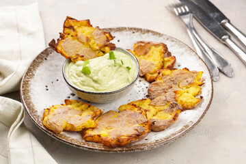 Trendy comfort snack crispy roasted crushed potatoes on a round plate with creamy avocado and garlic dip on a beige colored table cloth