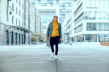 cool tough and handsome guy walk alone in city under skyscraper with grey coat and yellow sweater...
