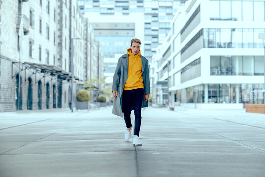 Cool Tough And Handsome Guy Walk Alone In City Under Skyscraper With Grey Coat And Yellow Sweater While Being Powerful And Show Fashion Outfit In City Life On Street