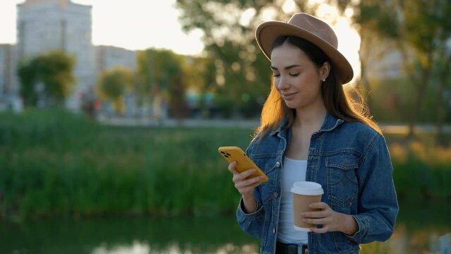 Beautiful caucasian woman using smartphone, wearing brown hat, walking near the local lake in the par holding eco coffee cup. People and technology concept