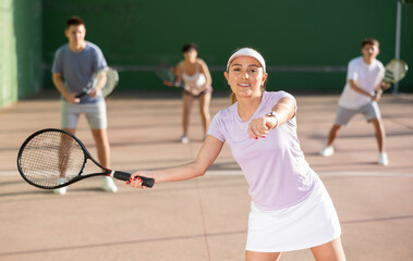 Sporty young Argentinian woman playing popular team match of frontenis at open-air fronton on summer day, ready to hit rubber ball with racquet