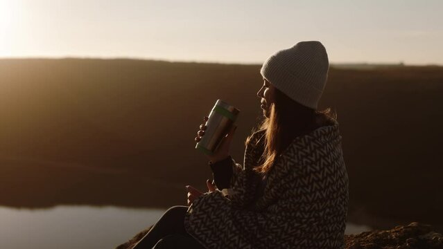 Beautiful tourist woman sitting high in the mountains on the rock of the canyon, wrapped in a blanket, drinking hot tea or coffee from a thermos. Offline satisfaction. Advertisement of mountain touris