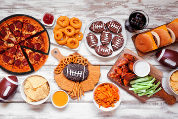 Super Bowl or football theme food table scene. Pizza, hamburgers, wings, snacks and sides. Top view on a white wood background.