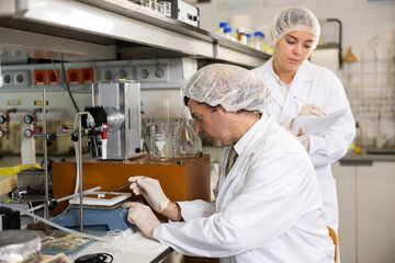 Serious middle-aged male scientist putting sample slide under microscope while female chemist taking notes in lab