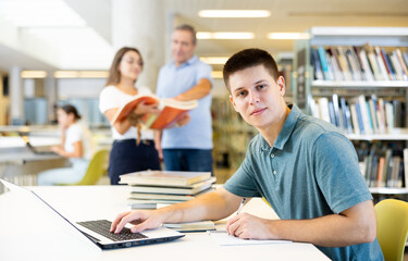 Fototapeta premium Concentrated young man spending time in library, reading books and making laptop. Self-education concept