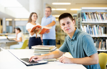 Concentrated young man spending time in library, reading books and making laptop. Self-education concept