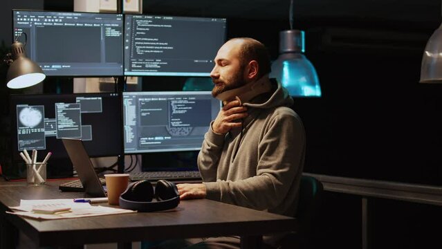 Software engineer wearing cervical collar at office, typing server code on multiple screens late at night. App developer with medical neck brace working with artificial intelligence. Handheld shot.