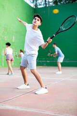 Sporty young Argentinian guy playing popular team game frontenis at open-air fronton court on summer day, ready to hit rubber ball with racquet