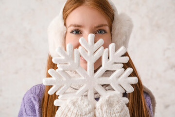Young redhead woman in warm earmuffs with big snowflake on light background, closeup