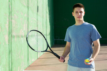 Portrait of positive caucasian boy standing on frontenis court, holding racket and ball