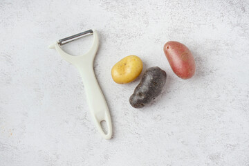 Different types of raw potatoes and peeler on light background