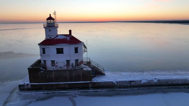 Isolated lighthouse blinking in winter on frozen lake at sunrise. Drone shot rising up with tilt down.