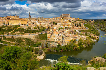 Fototapeta na wymiar View of historic area of Toledo city on banks of Tagus River overlooking belfry of Primatial Cathedral of Saint Mary and Alcazar fortress in cloudy spring day, Spain