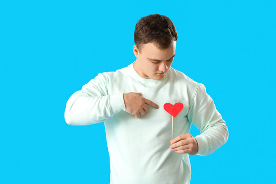 Young man pointing at paper heart for Valentine's Day on blue background