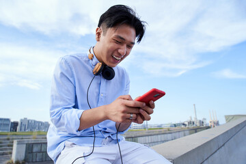 Young cheerful Asian man using mobile cell outdoors, texting and laughing. Happy Chinese boy typing...
