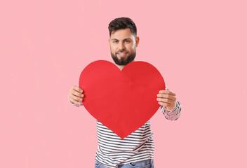 Young bearded man with big paper heart for Valentine's Day on pink background