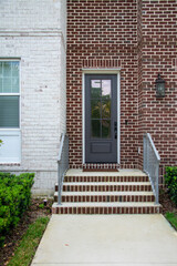 A single solid black and glass metal door in a brown and white brick building. The door handle is matt black. Green shrubs are next to the concrete steps. Greenery is reflecting in the door's glass. 